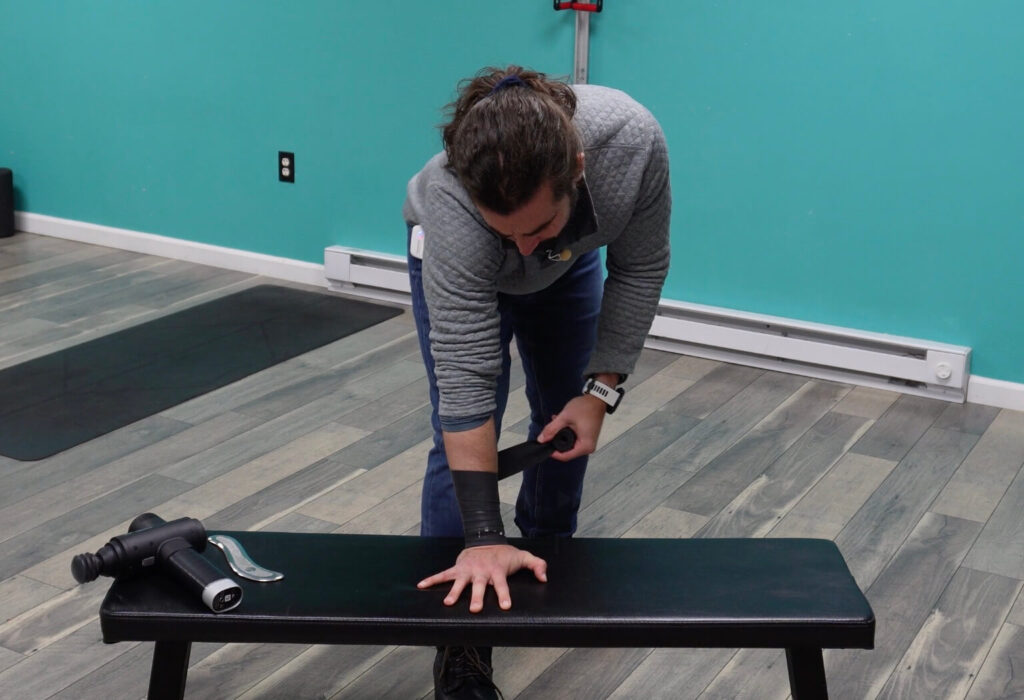 Dr. John with his right hand on a bench, wrist wrapped in muscle floss trying to break up scar tissue and improve mobility in the joint
